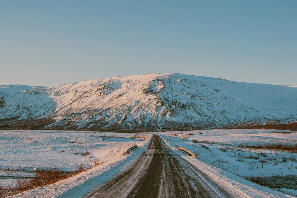 Carretera solitaria cubierta de nieve que conduce hacia montañas nevadas bajo un cielo despejado en Islandia.