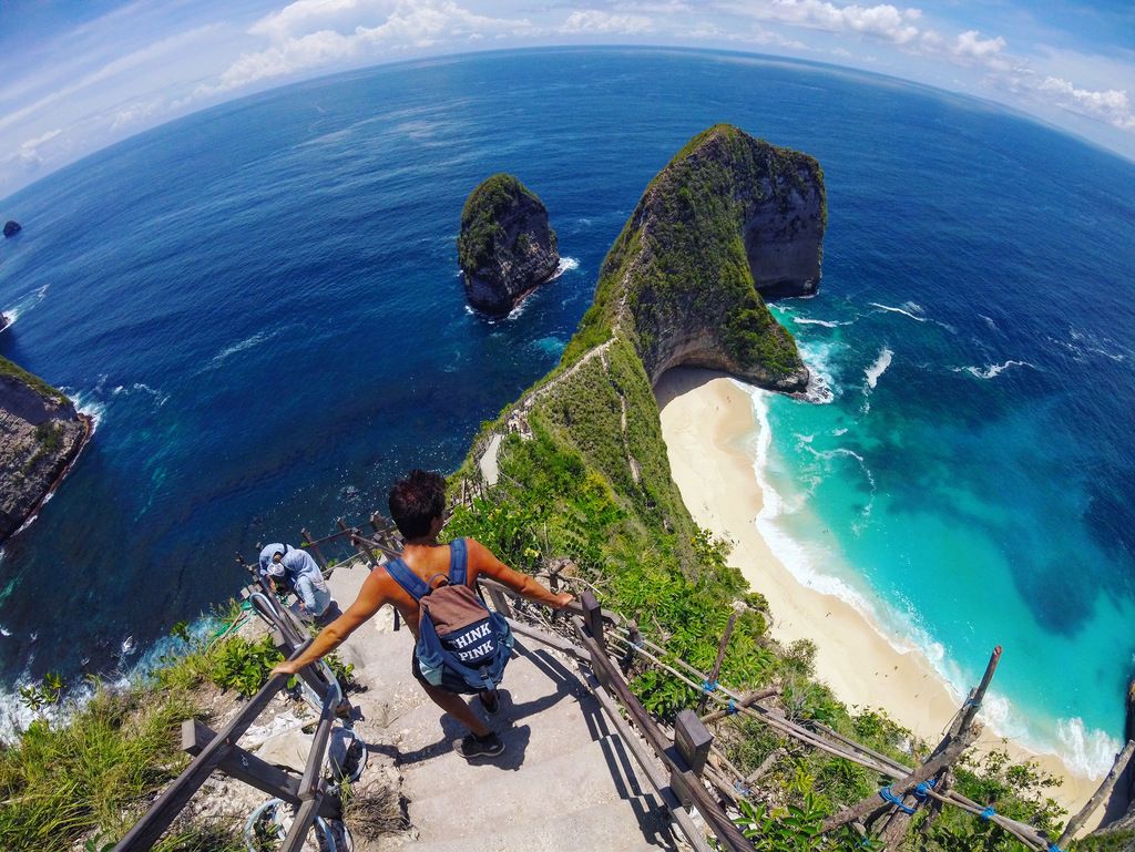 Turista descendiendo por las escaleras hacia la playa de Kelingking, en Nusa Penida, con vistas al mar turquesa y al acantilado en forma de dinosaurio.