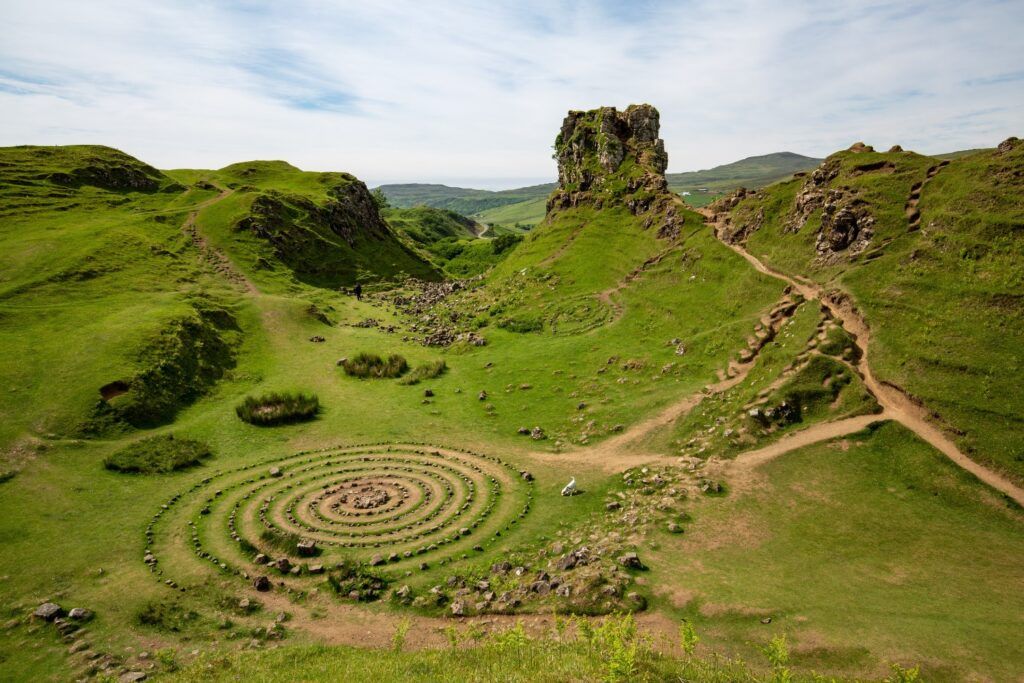 Vista aérea del Fairy Glen con colinas verdes, una formación rocosa distintiva (Castle Ewen) y un espiral de piedras en el suelo.