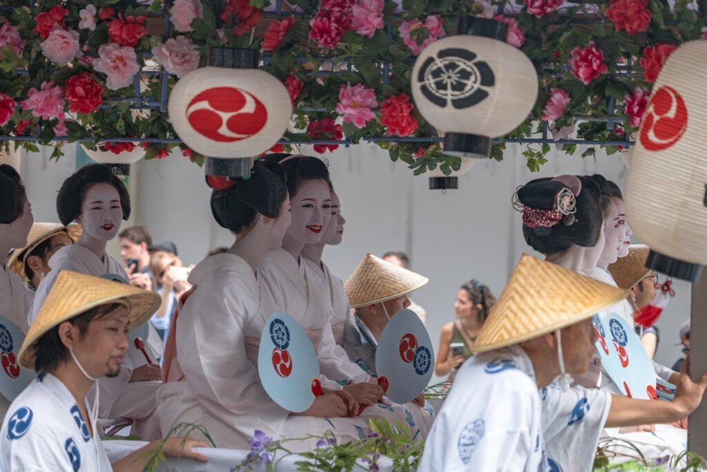 Grupo de mujeres con maquillaje de Geisha y hombres con sombreros de paja en una carroza de festival, bajo faroles de papel.
