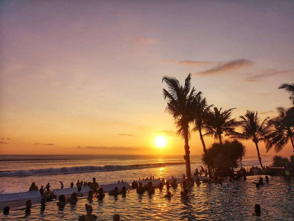 Puesta de sol tropical en una piscina o bar frente a la playa en Bali, con siluetas de palmeras y gente bañándose en el agua durante el atardecer.