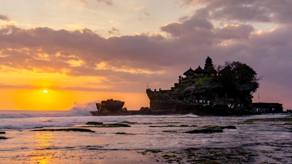 Santuario Tanah Lot en una roca azotada por las olas bajo un cielo de tonos naranjas al ocaso.