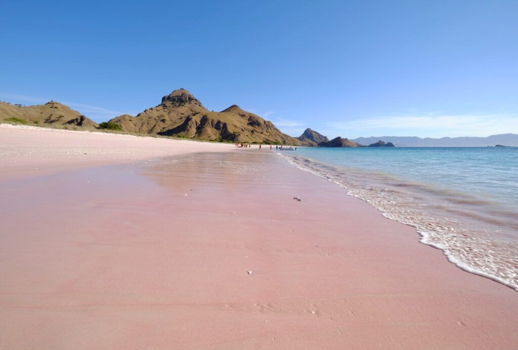 Vista de Pink Beach, con suaves olas de agua turquesa y montañas marrones de fondo bajo un cielo azul claro.
