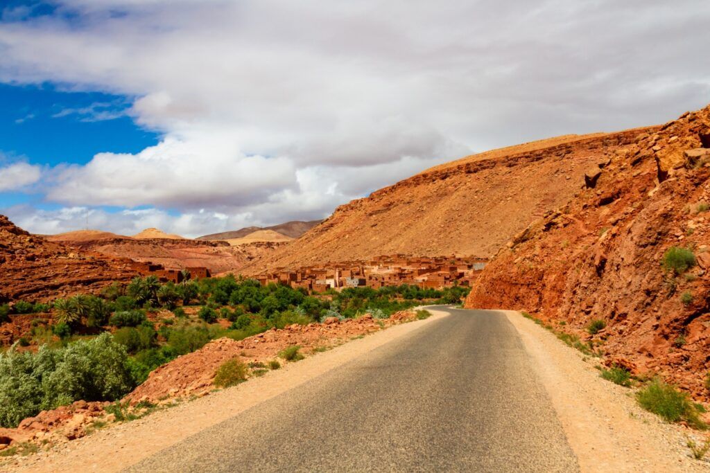 Carretera en el Atlas, con montañas rojas y un pueblo bereber de adobe entre el verde del valle.