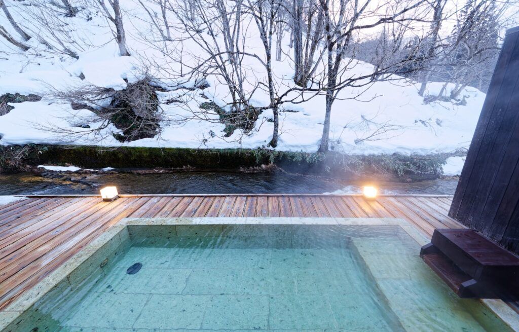 Rotenburo onsen exterior con nieve y vapor, junto a un río en Japón.