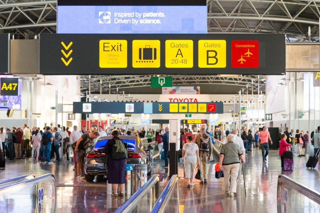 Vista interior y concurrida del área de llegadas del Aeropuerto de Bruselas, mostrando señales de salida, puertas de embarque y una gran multitud de viajeros.