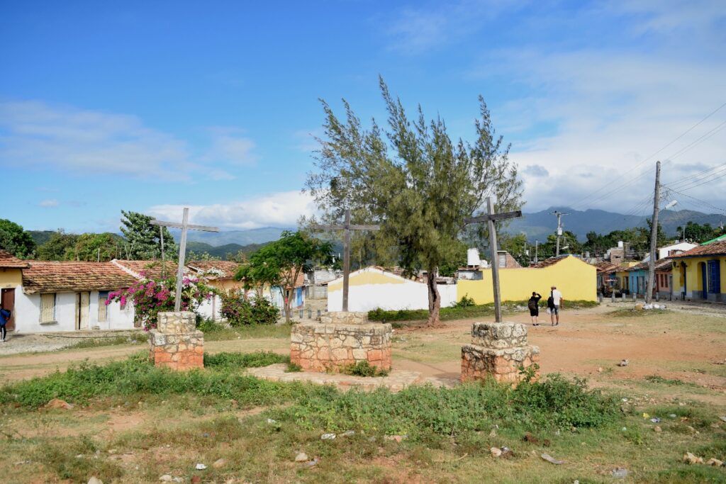 Cruces de madera sobre bases de piedra en una plaza rural de Trinidad, con casas de colores al fondo.