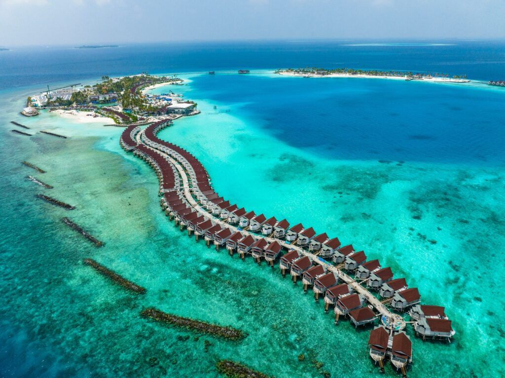 Une photographie d'un complexe hôtelier de luxe aux Maldives, avec une longue rangée de villas sur pilotis au toit brun reliées par une passerelle en bois.