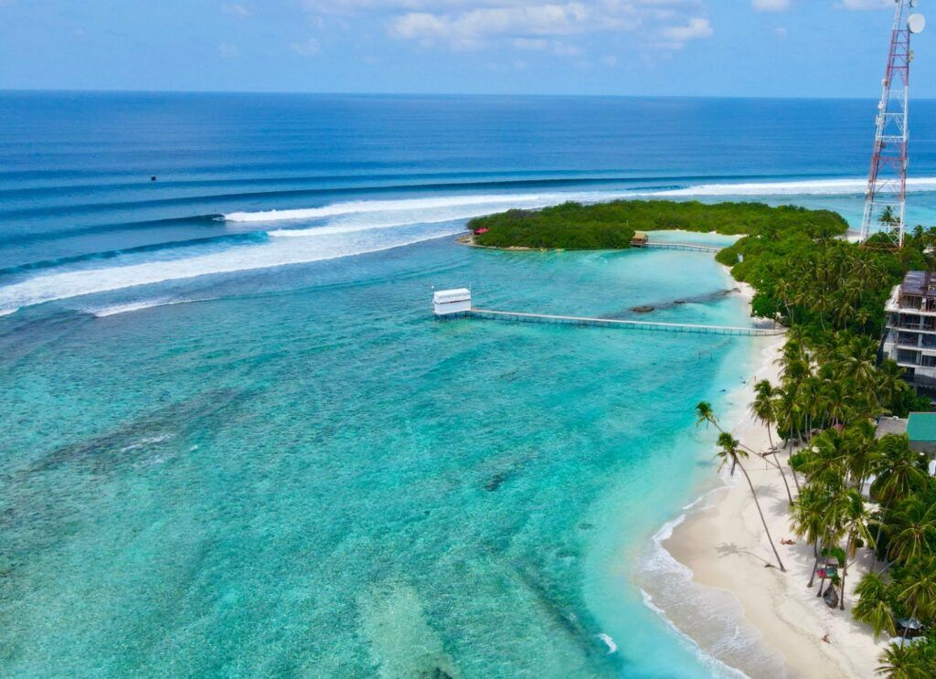Vue panoramique d'une plage de sable blanc avec des palmiers qui se balancent, des eaux cristallines et des vagues qui se brisent au loin dans l'océan.