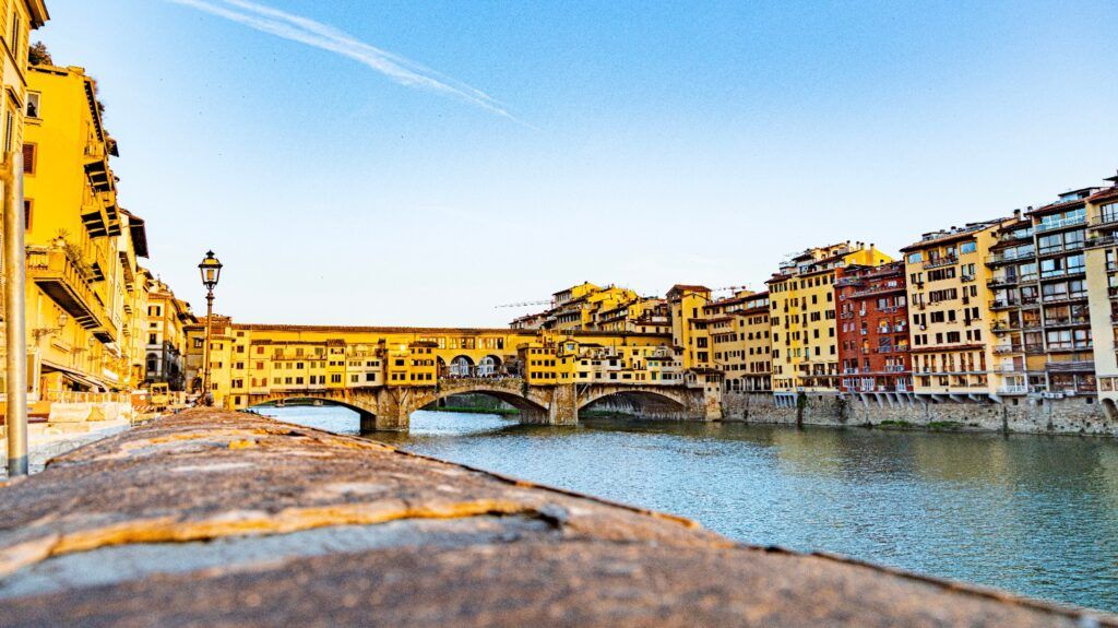 Vista lateral del Ponte Vecchio en Florencia sobre el río Arno, con sus casas colgantes bajo un cielo azul.