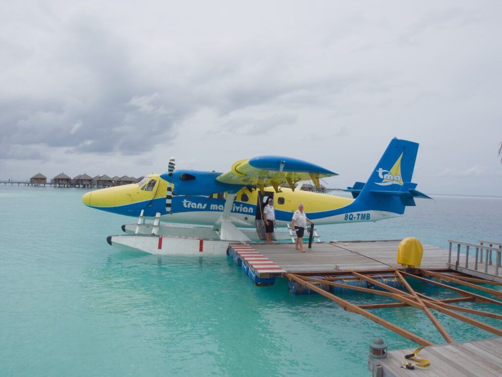 Un hydravion bleu et jaune de Trans Maldivian Airways amarré à un ponton flottant en bois sur des eaux turquoise transparentes.