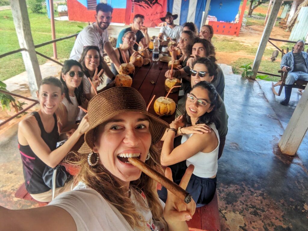 Un grupo de viajeros WeRoad sonrientes disfrutando de cocos frescos y cigarros juntos en una larga mesa de madera en Cuba.