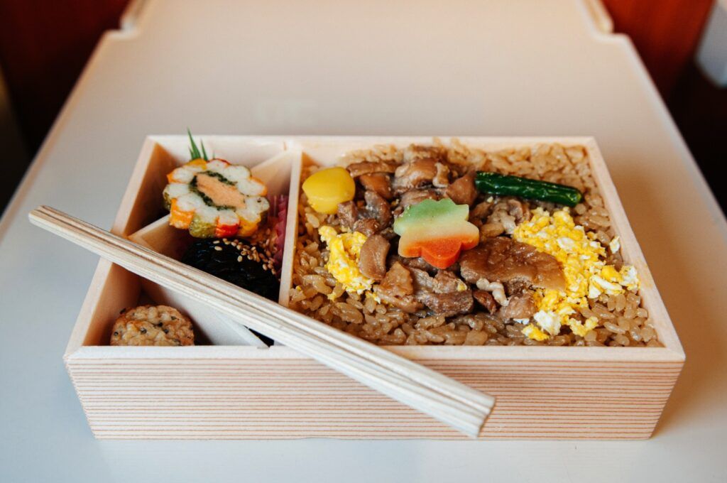 Un ekiben japonés de primavera en una caja de madera, con arroz, carne, huevo y decoraciones vegetales coloridas.