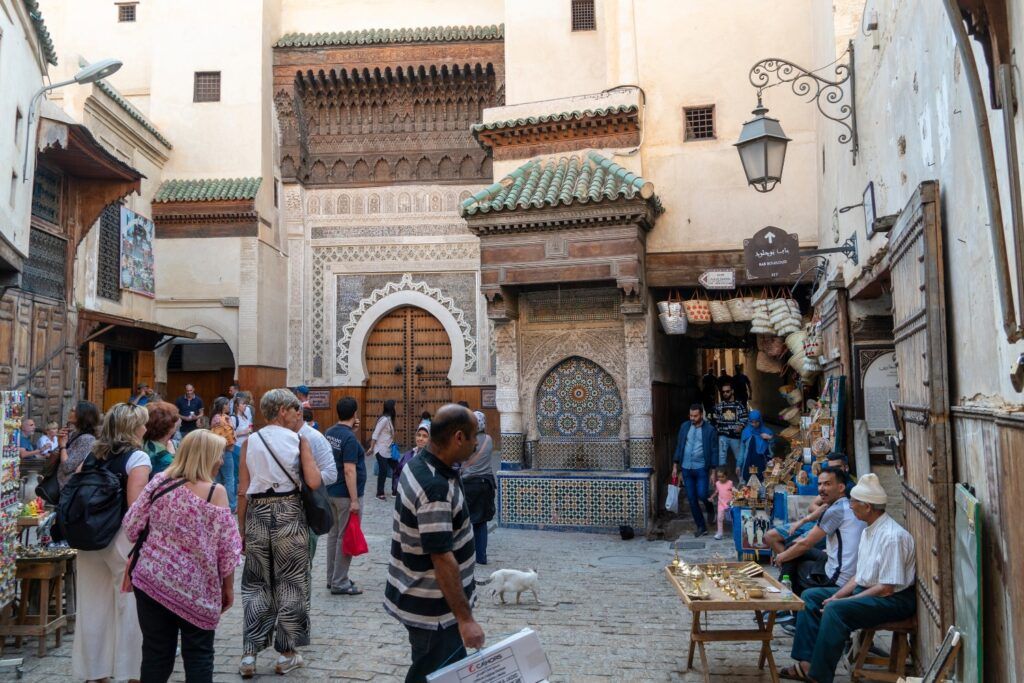 Calle concurrida en la medina de Fez con turistas caminando frente a una fuente tradicional de mosaicos y edificios antiguos con detalles de madera tallada.