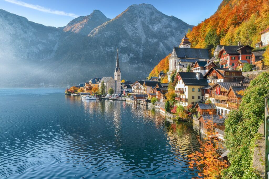 El pintoresco pueblo de Hallstatt a orillas de un lago, con casas tradicionales de madera y una iglesia rodeadas de montañas alpinas y colores otoñales.