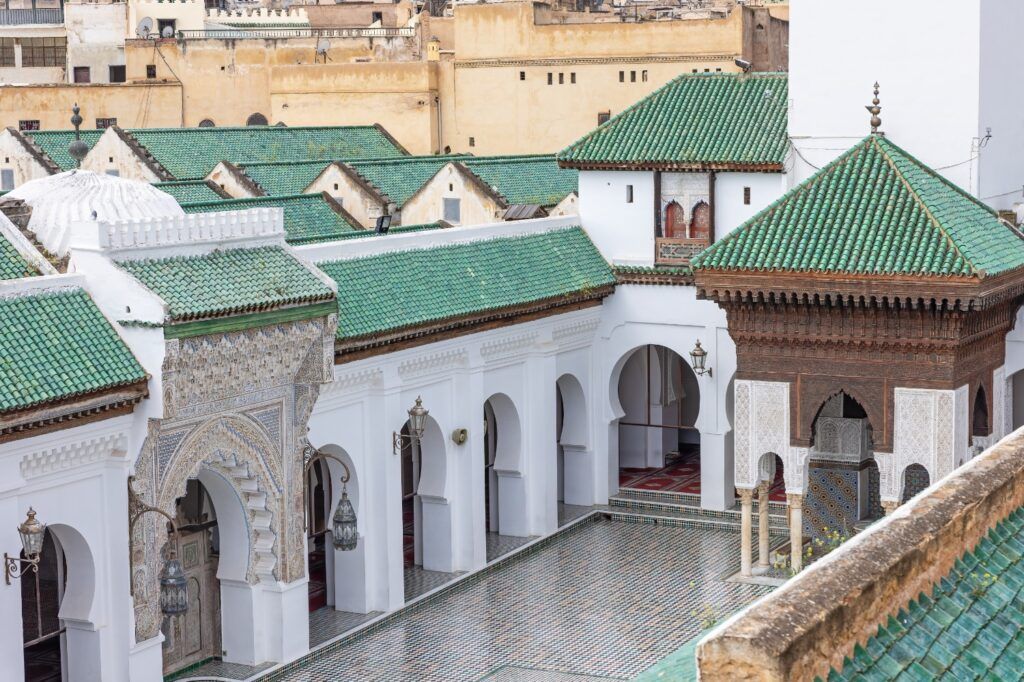 Vista aérea del patio interior de la Mezquita Al-Qarawiyyin en Fez, con sus característicos tejados de tejas verdes, arcos blancos decorados y pavimentos de azulejos detallados.