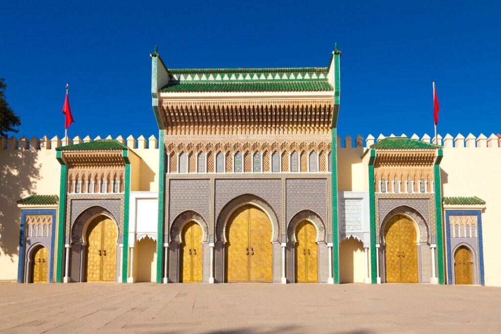 Fachada del Palacio Real de Fez con sus siete imponentes puertas de bronce dorado, decoradas con intrincados mosaicos de azulejos y techos de tejas verdes bajo un cielo azul despejado.